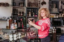 Feliz pelo rizado mujer rubia vendedora barman con copa de vidrio con vino mientras trabaja en la tienda de alimentos delicatessen locales - foto de stock