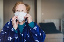 Seniorin mit roten Haaren in blauem Gewand und medizinischer Maske blickt im Krankenhauszimmer in die Kamera — Stockfoto