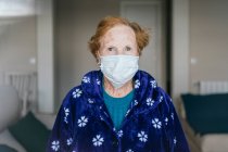 Mujer mayor con el pelo rojo en bata azul y máscara médica mirando a la cámara mientras está de pie en la habitación del hospital - foto de stock