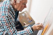 Вид збоку на культуру старший чоловічий художник у повсякденному одязі, малюнок з пензлем на папері на мольберті, що тримає палітру з фарбами у творчій майстерні — стокове фото