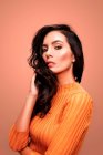 Charming confident young brunette female with long wavy hair and perfect makeup wearing casual orange sweater looking at camera while standing against pink background — Stock Photo