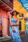 Jovem calmo no elegante vestido de verão azul e chapéu olhando para a câmera enquanto estava perto do edifício envelhecido com paredes coloridas na rua da cidade — Fotografia de Stock