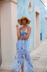 Gorgeous young female in beautiful sundress and straw hat looking at camera while standing near aged building with blue walls on street — Stock Photo