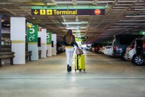 Adolescent en vêtements décontractés avec les cheveux longs marchant dans le parking et portant une valise jaune dans le bâtiment de l'aéroport — Photo de stock