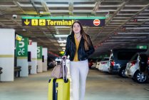Young positive just arrived teenager in casual clothes with long hair walking in parking and carrying yellow suitcase in airport building — Stock Photo