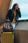 Adolescente em roupas casuais com mala e bolsa em pé pela janela inclinada na parede e falando no smartphone no aeroporto — Fotografia de Stock