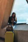 Adolescente en ropa casual con maleta y bolsa de pie junto a la ventana apoyada en la pared y hablando en el teléfono inteligente en el aeropuerto - foto de stock