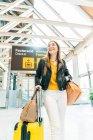 Elegante adolescente sonriente con gorra amarilla chaqueta de cuero negro y blusa amarilla de pie con maleta en la moderna terminal del aeropuerto y mirando a la cámara - foto de stock
