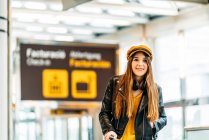 Stylish smiling teenager in yellow cap black leather jacket and yellow blouse with headphones standing with suitcase in modern airport terminal and looking at camera — Stock Photo