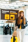 Stylish smiling teenager in yellow cap black leather jacket and yellow blouse with headphones standing with suitcase in modern airport terminal and looking at camera — Stock Photo