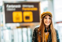 Teenager in yellow cap black leather jacket and yellow blouse with headphones standing with suitcase in modern airport terminal and looking at camera — Stock Photo