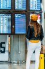 Back view of teenager in trendy outfit with baggage standing near schedule and checking departure time on digital display while waiting for flight at airport terminal — Stock Photo