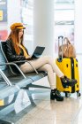 Seitenansicht eines ernsten Teenagers in stylischer Kleidung, der mit gekreuzten Beinen auf einer Bank sitzt und seine Freizeit mit Laptop verbringt, während er auf das Boarding im Flughafen wartet — Stockfoto