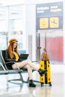 Side view of serious teenager in stylish clothes sitting on bench with legs crossed with luggage and spending free time using laptop while waiting for boarding in airport — Stock Photo