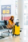 Side view of serious teenager in stylish clothes sitting on bench with legs crossed with luggage and spending free time using laptop while waiting for boarding in airport — Stock Photo