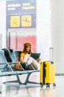 Вид сбоку серьезного подростка в стильной одежде, сидящего на скамейке со скрещенными ногами и проводящего свободное время с ноутбуком в ожидании посадки в аэропорту — стоковое фото