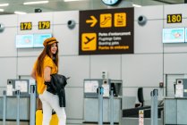 Full length of joyful teenager in fashionable outfit with luggage and passport in back pocket looking at camera and waving goodbye while standing next to check in counter at airport — Stock Photo