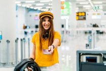 Alegre adolescente sonriente en colorida ropa elegante y auriculares que dan pasaporte y mirando a la cámara mientras está de pie en el mostrador en la terminal del aeropuerto - foto de stock