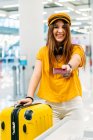 Adolescent souriant joyeux dans des vêtements élégants colorés et écouteurs donnant passeport et regardant la caméra tout en se tenant au comptoir d'enregistrement dans le terminal de l'aéroport — Photo de stock