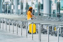 Corpo inteiro de adolescente elegante irreconhecível em roupa colorida com mala de plástico amarelo recebendo carrinho de bagagem enquanto estava perto do edifício do aeroporto — Fotografia de Stock