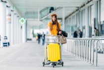 Smiling teenager in casual outfit and headphones with leather jacket bag and passport in hand looking at camera and carrying luggage cart near modern airport building — Stock Photo