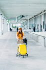 Smiling teenager in casual outfit and headphones with leather jacket bag and passport in hand looking at camera and carrying luggage cart near modern airport building — Stock Photo