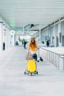 Lächelnder Teenager in lässigem Outfit und Kopfhörer mit Lederjacke und Reisepass in der Hand, der in die Kamera blickt und in der Nähe des modernen Flughafengebäudes einen Gepäckwagen trägt — Stockfoto
