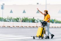 Ein fröhlicher Teenager in gelber Mütze mit Kopfhörern und vollem Gepäckwagen, der an warmen Sommertagen in der Nähe der Straße vor dem großen Flughafenterminal steht und ein Taxi auffängt. — Stockfoto