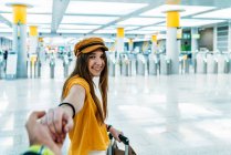 Молодой улыбчивый подросток в стильном наряде ведет кого-то вручную и смотрит в камеру на пути в комнату ожидания с чемоданом в терминале аэропорта — стоковое фото