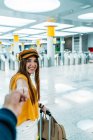 Молодий усміхнений підліток у стильному вбранні веде когось вручну і дивиться на камеру на шляху до кімнати очікування з валізою в терміналі аеропорту — стокове фото