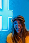 Щасливий молодий підліток в жовтій сорочці і модна шапка робить смішну похмурість і показує язик на синій стіні з неоновим знаком медичного хреста — стокове фото
