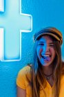 Felice giovane adolescente in t shirt gialla e berretto alla moda facendo smorfia divertente e mostrando la lingua contro il muro blu con neon segno di croce medica — Foto stock
