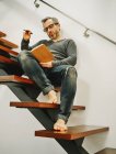 Low angle of mature man in eyeglasses relaxing with cup of coffee on wooden steps in contemporary apartment and writing plans in notebook — Stock Photo