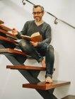 Low angle of happy mature man in eyeglasses relaxing with cup of coffee on wooden steps in contemporary apartment and writing plans in notebook — Stock Photo