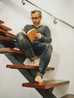 Низкий угол зрелого мужчины в очках расслабляющий с чашкой кофе на деревянных ступеньках в современной квартире и написание планов в блокноте — стоковое фото
