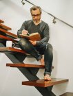 Angolo basso di uomo maturo in occhiali da vista rilassante con tazza di caffè su gradini di legno in appartamento contemporaneo e piani di scrittura nel quaderno — Foto stock