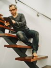 Счастливый человек в повседневной одежде сидит на деревянной лестнице с помощью планшета во время отдыха в современном доме — стоковое фото