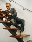 Uomo felice in abbigliamento casual seduto su scale di legno utilizzando tablet durante il riposo in casa moderna — Foto stock