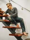 Low angle of mature man in eyeglasses relaxing with cup of coffee on wooden steps in contemporary apartment and writing plans in notebook — Stock Photo