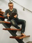 Homem feliz em roupas casuais sentado em escadas de madeira usando tablet durante o descanso em casa moderna — Fotografia de Stock