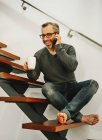 Lachender Mann in lässiger Kleidung sitzt auf Holztreppen und plaudert mit dem Handy, während er in der modernen Wohnung ein Heißgetränk genießt — Stockfoto