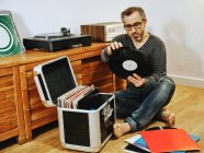 Stylish man wearing casual clothes and eyeglasses sitting on wooden floor and picking vinyl record while enjoying weekend at home — Stock Photo