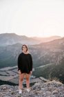 Full body cheerful female tourist in casual black clothes standing on stony cliff in mountains and smiling while looking at camera during sunset — Stock Photo