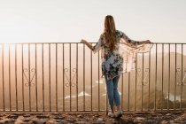 Visão traseira de turista feminino em roupas casuais em pé perto de cerca de metal e admirando a vista do pôr do sol durante a caminhada no dia de verão — Fotografia de Stock