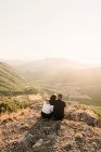 Back view of romantic couple of tourists in casual clothes sitting on stone edge of cliff embracing and enjoying picturesque landscape during sunny day — Stock Photo
