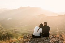 Вид на романтическую пару туристов в повседневной одежде, сидящих на каменном краю скалы, обнимающих и наслаждающихся живописным пейзажем в солнечный день — стоковое фото