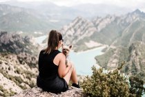 Back view of anonymous female tourist in casual black clothes and sunglasses sitting with dog on rocky hill and admiring picturesque scenery in summer — Stock Photo
