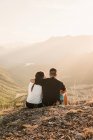 Вид на романтическую пару туристов в повседневной одежде, сидящих на каменном краю скалы, обнимающих и наслаждающихся живописным пейзажем в солнечный день — стоковое фото