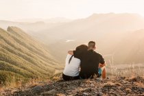 Visão traseira de casal romântico de turistas em roupas casuais sentado na borda de pedra do penhasco abraçando e desfrutando de paisagem pitoresca durante o dia ensolarado — Fotografia de Stock