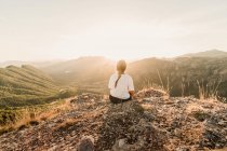 Vista posteriore di anonimo turista donna in abiti casual godendo paesaggio pittoresco mentre seduto su un terreno pietroso nella giornata di sole in estate — Foto stock